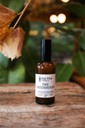 Bottle of 'The Greenhouse' essential oil room spray by Salt Folk Trading Co. on a wooden surface with leaves in the background.