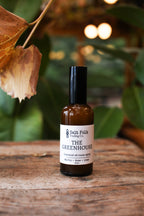 Bottle of 'The Greenhouse' essential oil room spray by Salt Folk Trading Co. on a wooden surface with leaves in the background.