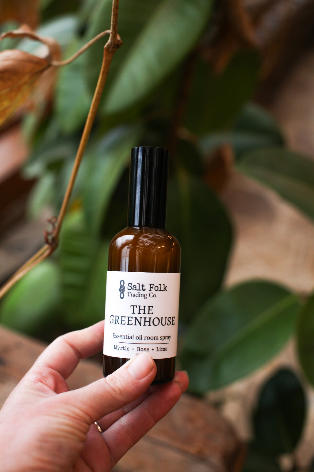Hand holding a bottle labeled 'The Greenhouse' by Salt Folk Trading Co. with green leaves in the background.