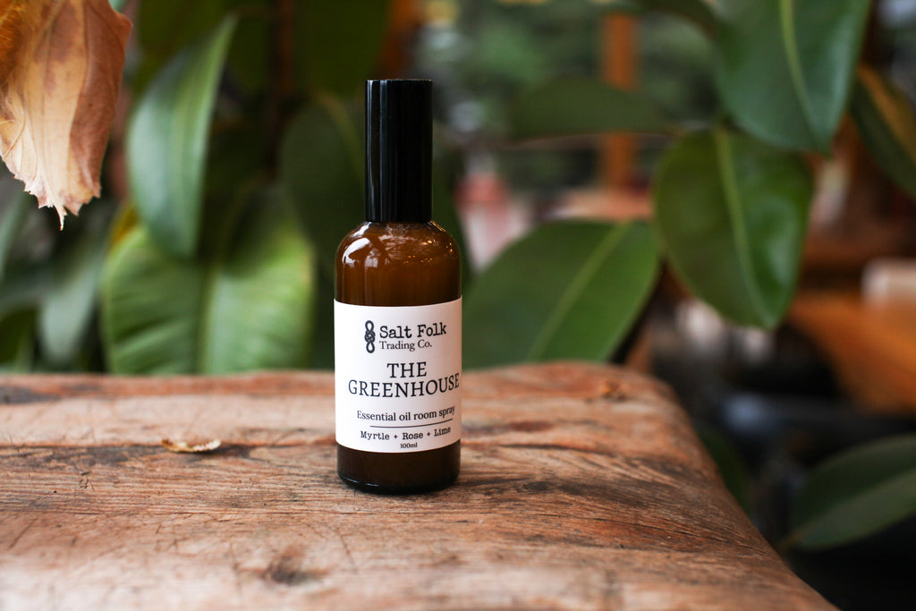 Bottle of 'The Greenhouse' essential oil on a wooden surface with plants in the background