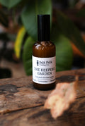 Bottle of 'The Keeper's Garden' essential oil room spray by Salt Folk Trading Co. on a wooden surface with green leaves in the background.